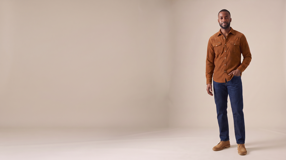 Man in a rust corduroy western shirt standing on a plain stone backdrop